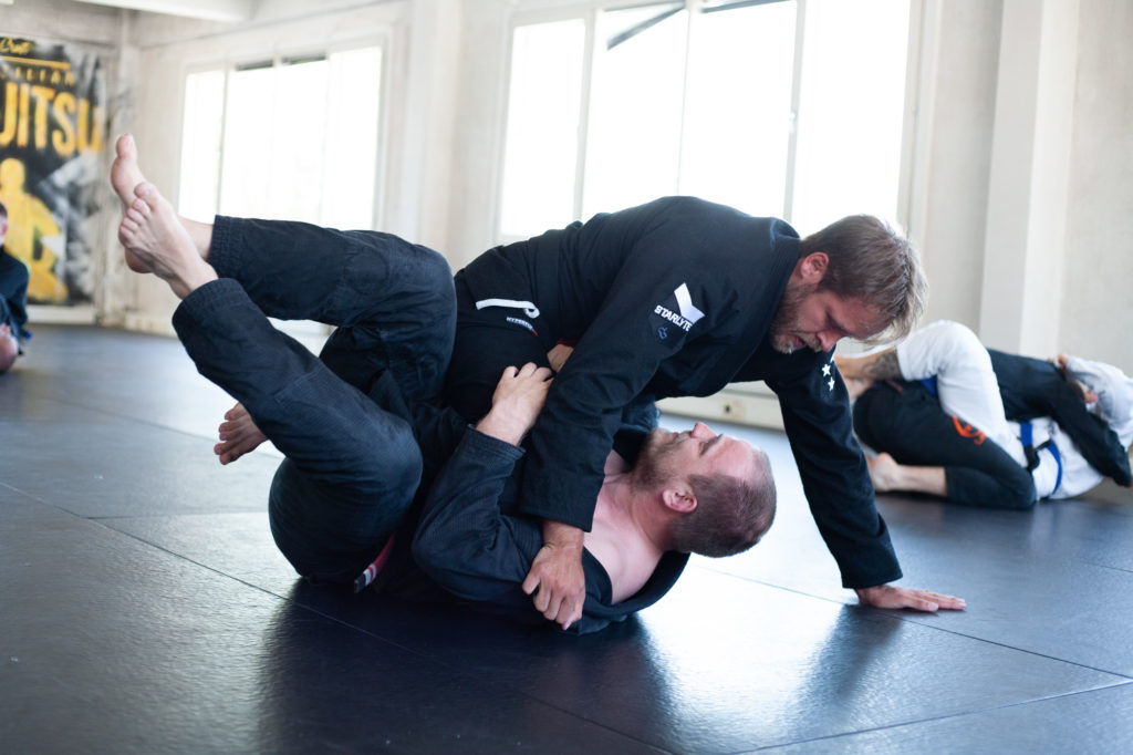 Two large, masculine grapplers. The one on top is posting his arm on the mat.