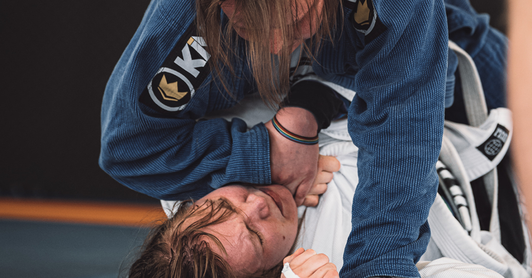 A jiu jitsu match, a larger peraon with long hair is being controlled by a smaller person sitting on them.