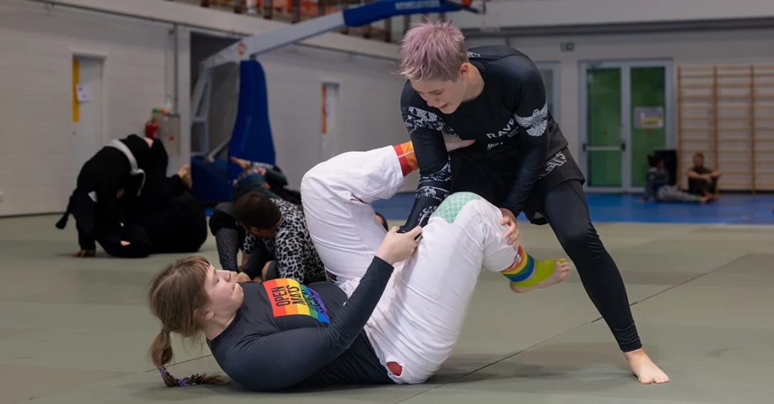 Two people grappling in no-gi. One on of them has short pink hair, the other wears an Open Mats Open Minds rash guard.